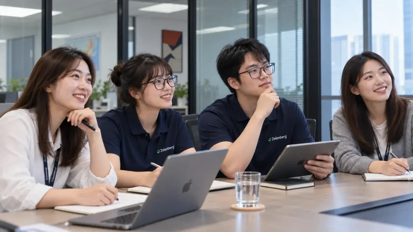 Four colleagues at a meeting table with a laptop and a tablet, listening and smiling