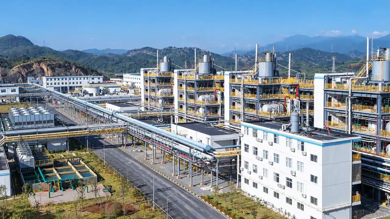 A processing plant seen from above: towers, pipework and a white office block, with hills behind