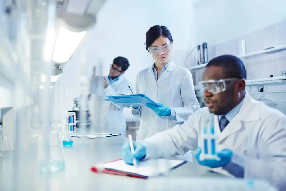 Three people in white coats and safety glasses working at a laboratory bench; one holds a clipboard